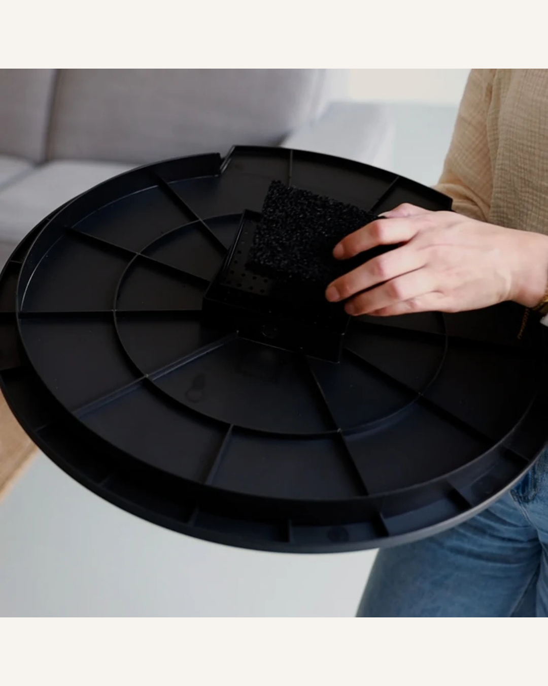 Hand holding a small black carbon filter pad above a large circular black plastic tray with segmented sections.