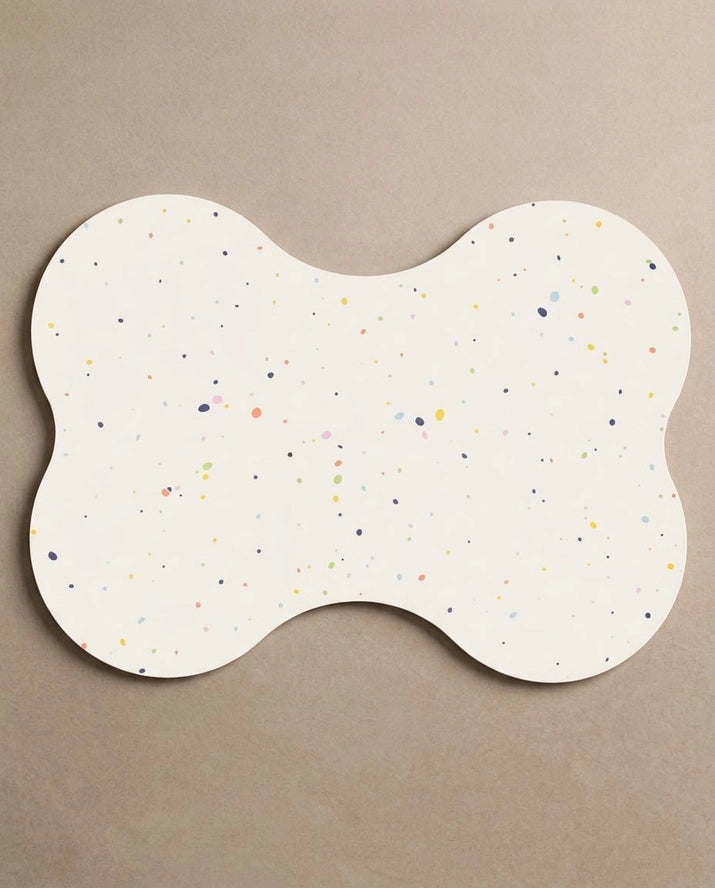 Bone-shaped dog bowl placemat in white with multicolored speckles on a beige background.