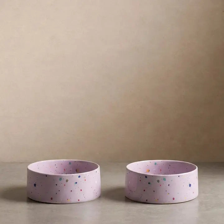 Two round pink ceramic pet bowls with colorful speckles on a smooth surface.