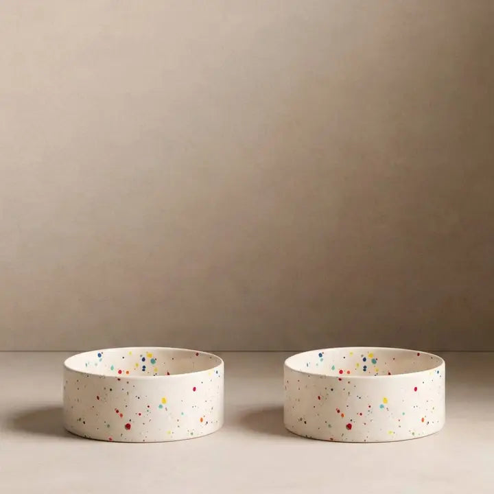 Set of two round white ceramic pet bowls with colorful confetti speckles and shallow sides.
